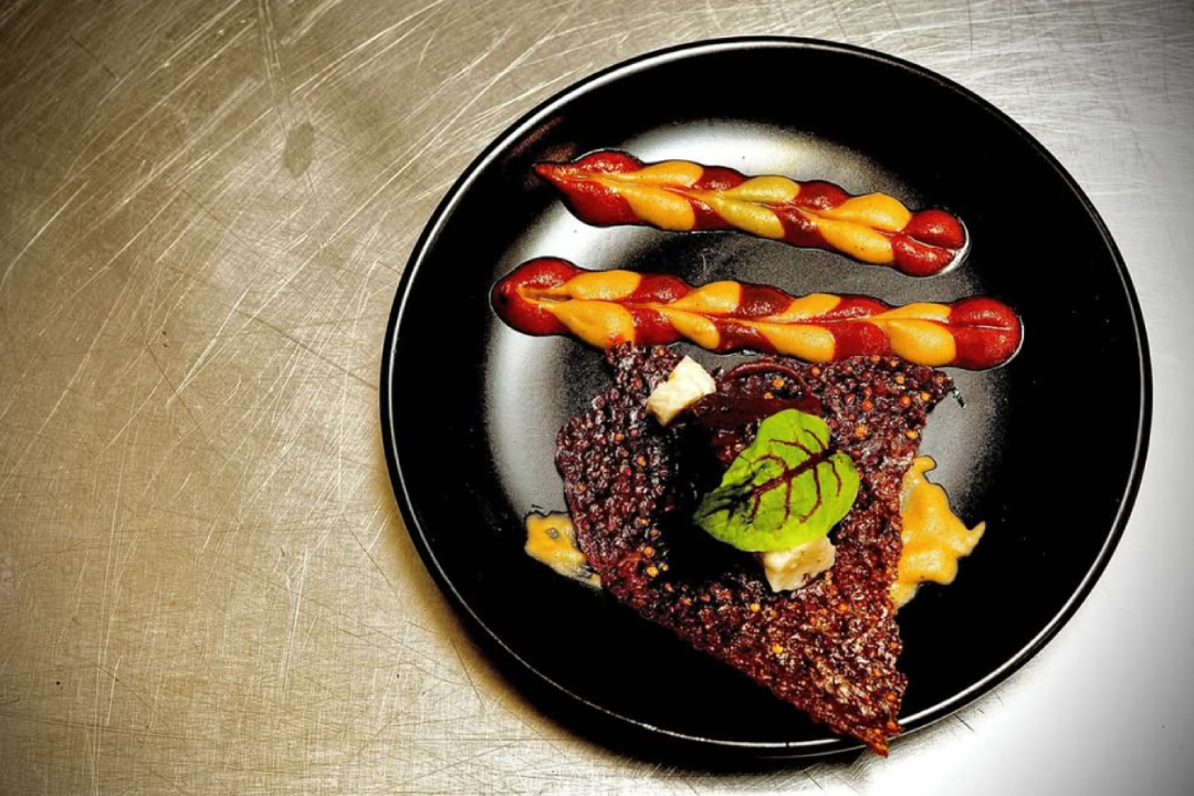 An inviting outdoor patio space illuminated by string lights draped across trees and shrubs. In the foreground, three round cocktail tables with white tablecloths are topped with lit candles. In the background, a long, curved built-in bench with pillows in shades of white, orange, and gray provides additional seating. The patio floor is made of light-colored pavers, and the surrounding area is landscaped with various green trees and bushes, creating a serene evening ambiance.