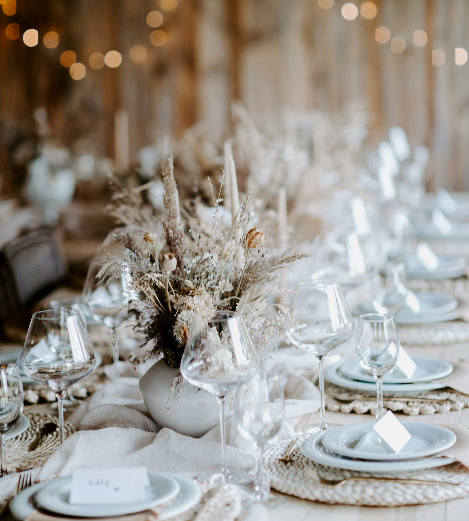 A beautifully set wedding or event table with a rustic, bohemian theme. It features a centerpiece of dried pampas grass and flowers in a white vase, surrounded by clear wine glasses, light-colored plates on woven placemats, and soft fabric runners. Blurry fairy lights are visible in the background.