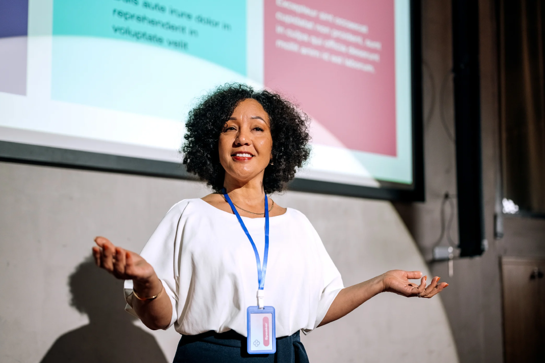 A speaker on a stage in a ballroom, presenting in front of a large screen that displays "Product School" and an image related to "Product Manager." The stage is lit with blue and white lights, and a banner for "Product School Silicon Valley" is visible.